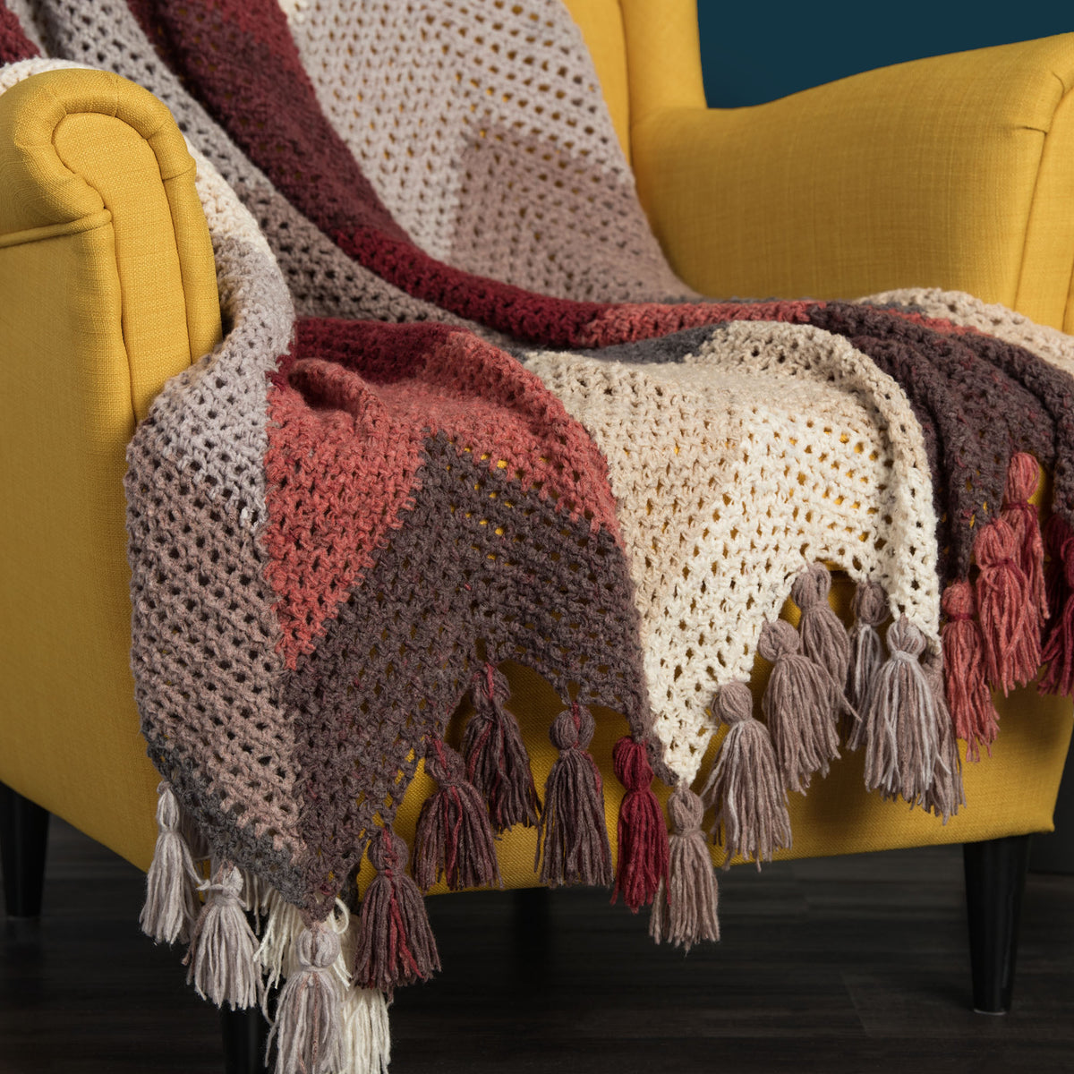 A chunky knit throw blanket with a chevron pattern in beige, rust red, brown, and dark gray, finished with fringed tassels.