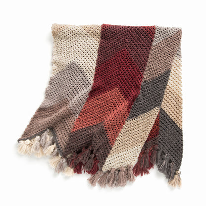 Caron Cloud Chevron Stripes Crochet Blanket A chunky knit throw blanket with a chevron pattern in beige, rust red, brown, and dark gray, finished with fringed tassels.