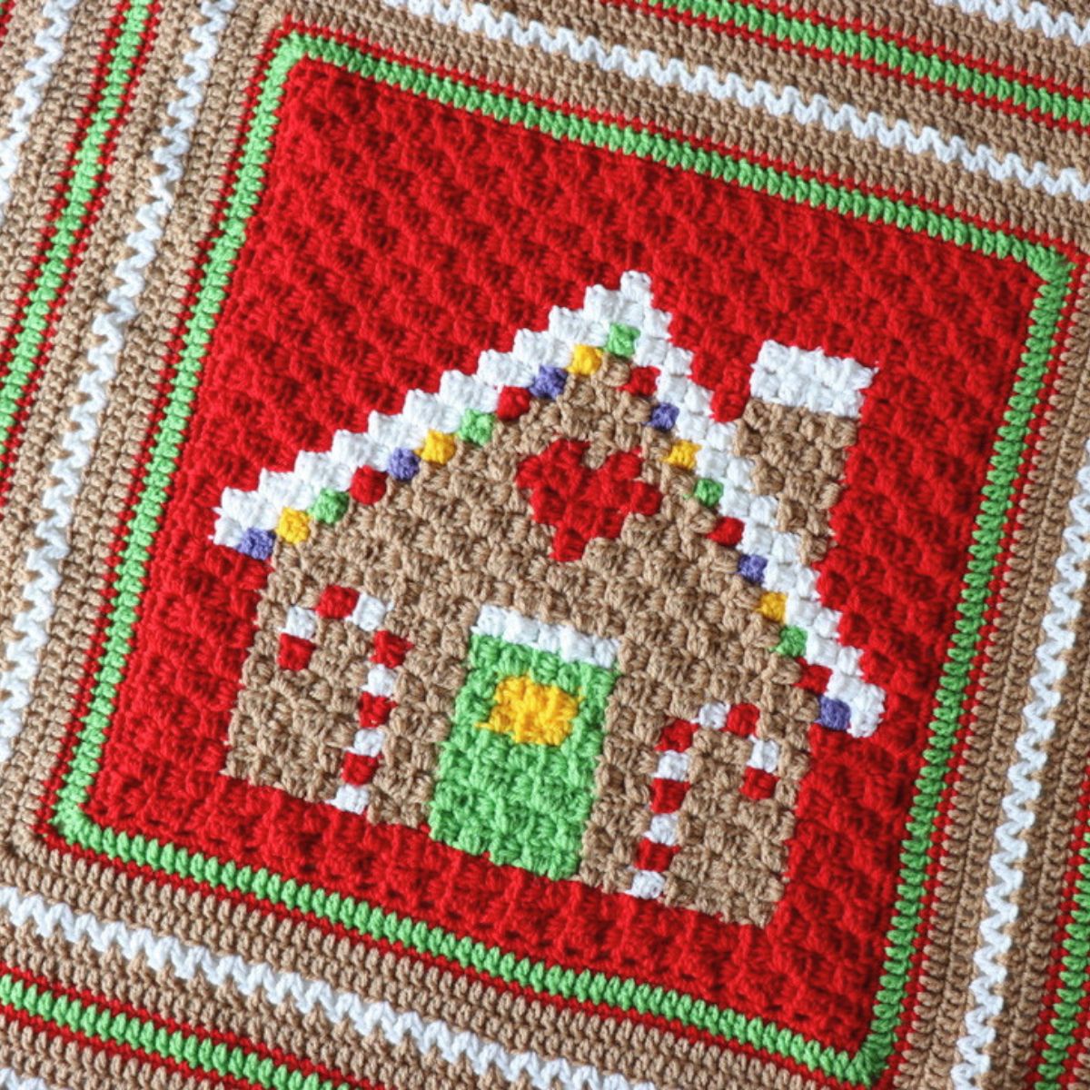 A handmade crocheted blanket featuring a gingerbread house design, draped over a chair in front of a shelf filled with colorful yarn skeins.