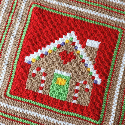 Red Heart Gingerbread House Blanket A handmade crocheted blanket featuring a gingerbread house design, draped over a chair in front of a shelf filled with colorful yarn skeins.