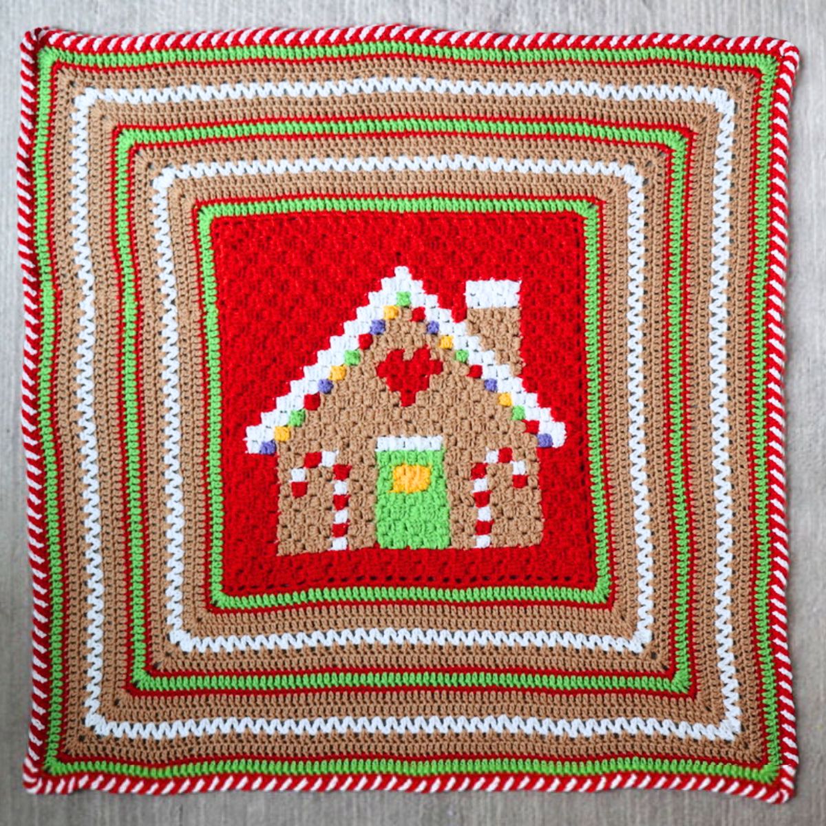 A handmade crocheted blanket featuring a gingerbread house design, draped over a chair in front of a shelf filled with colorful yarn skeins.