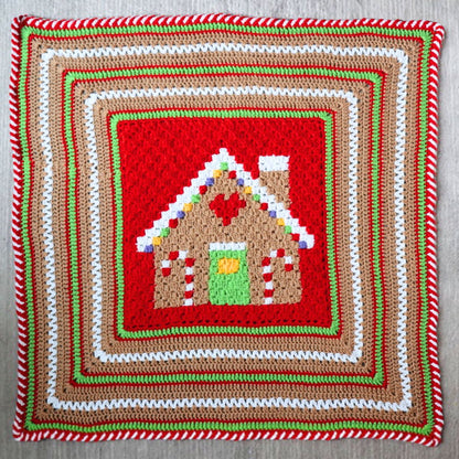 Red Heart Gingerbread House Blanket A handmade crocheted blanket featuring a gingerbread house design, draped over a chair in front of a shelf filled with colorful yarn skeins.