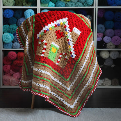 Red Heart Gingerbread House Blanket A handmade crocheted blanket featuring a gingerbread house design, draped over a chair in front of a shelf filled with colorful yarn skeins.
