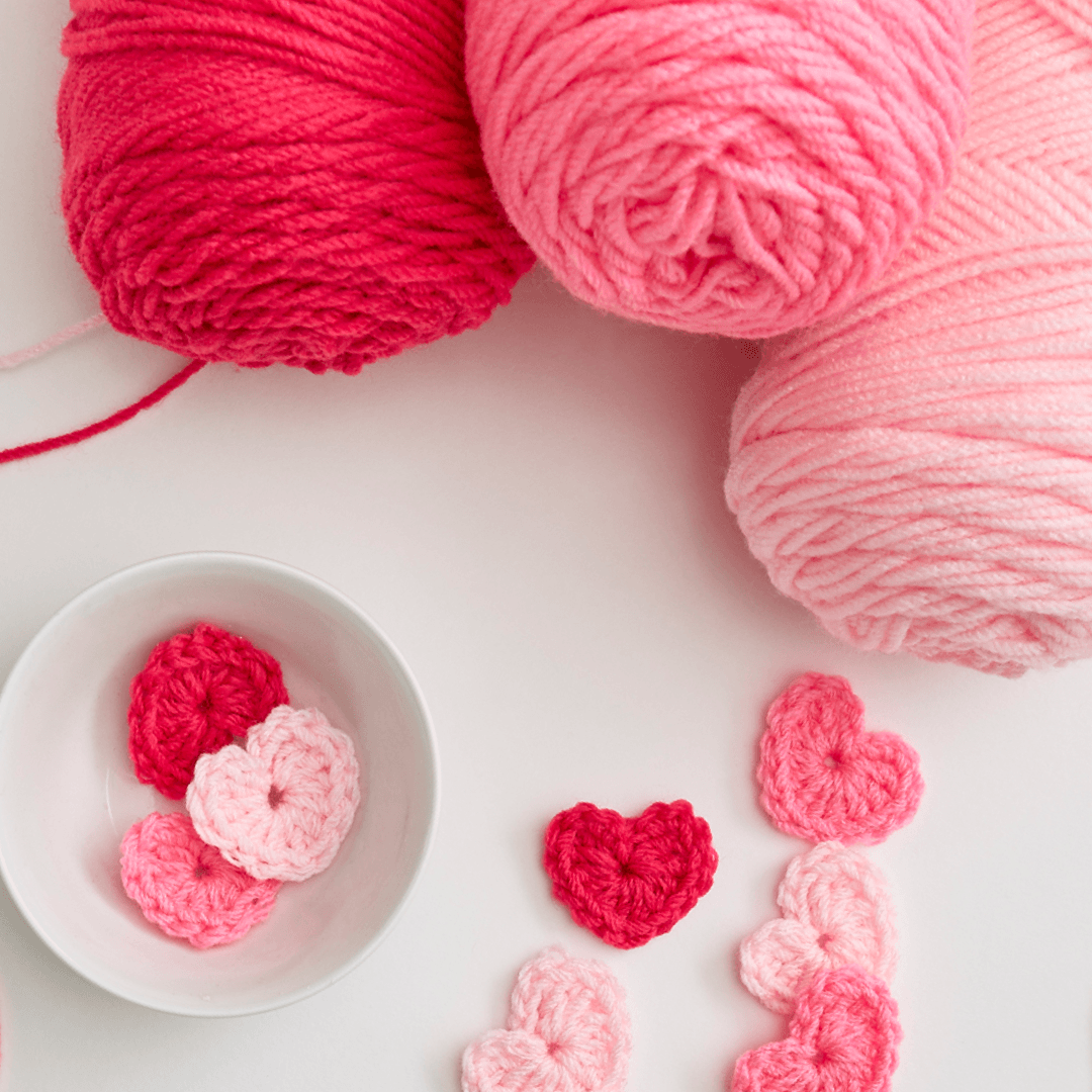 Red Heart brand's yarns & crocheted heart applique on a white background.