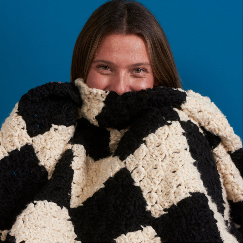 A woman partially covering her face with a crochet checkerboard blanket made from Yarnspirations yarns.